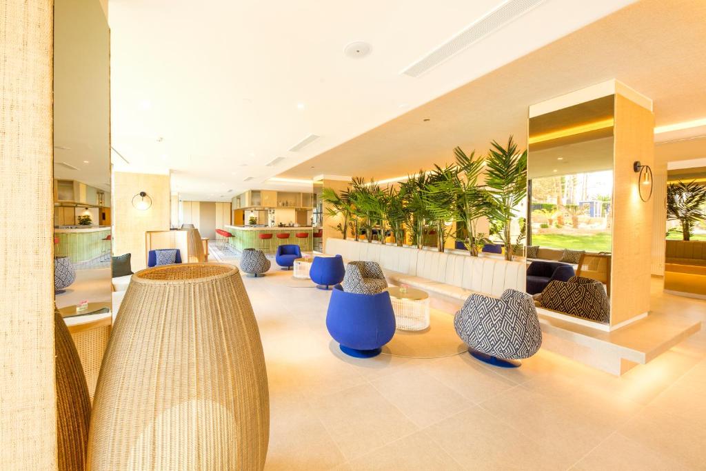 Mūsdienīgs viesnīcas interjers ar stilīgām zālēm, ko rotā augu kompozīcijas un krāsaini sēdekļi. Telpa ir gaiša un plaša, radot viesmīlīgu atmosfēru.