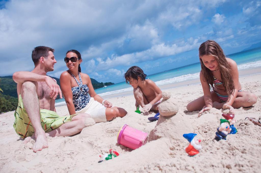 Ģimenes laiks pludmalē, kur bērni spēlējas ar smiltīm un veido smilšu pilis, kamēr pieaugušie sarunājas un atpūšas. Skaista jūras ainava un saulaina debesis papildina šo prieka mirkli.