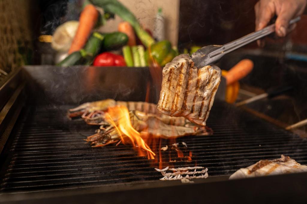 Grilēšanas process ar gaļu un dārzeņiem, kas izdala tvaikus un liesmas. Dārzeņi ir redzami fonā, radot gardējo atmosfēru.