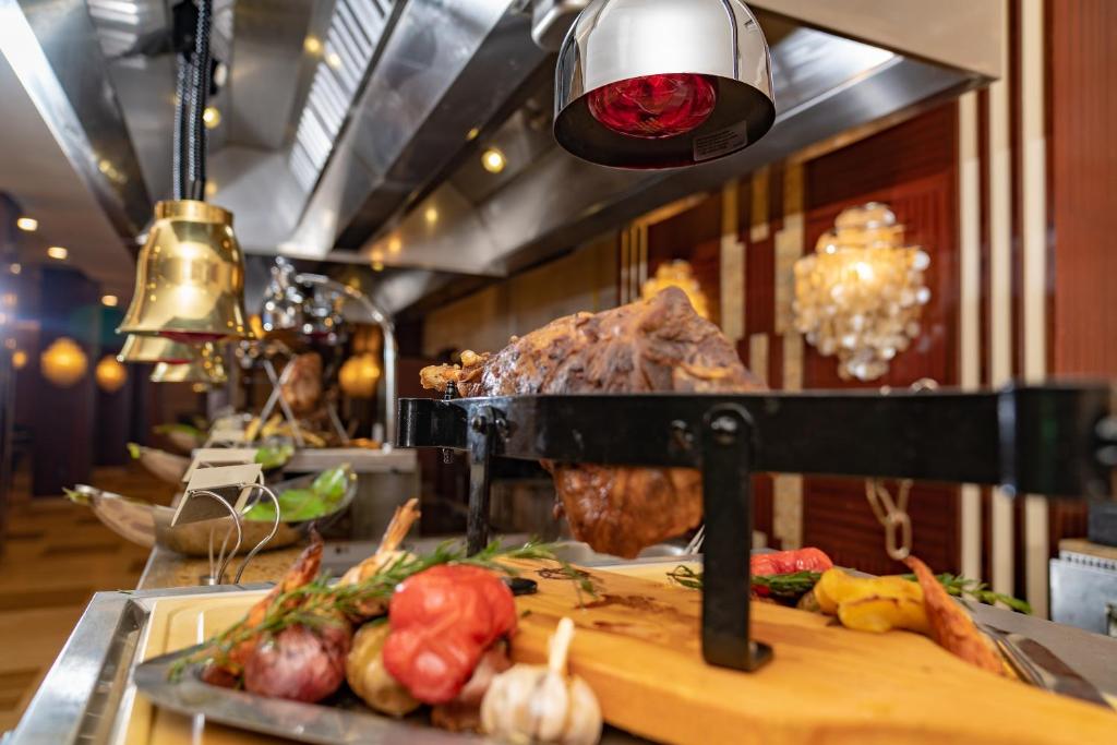 Eleganta restorāna bufete ar dažādiem gaļas ēdieniem un dārzeņiem, kas tiek pasniegti uz melnas plāksnes. Fonā redzami spoži gaismas elementi un moderns interjers.