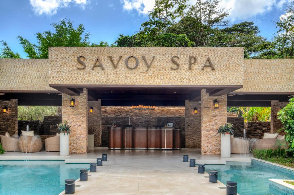 Savoy Spa ieejas zona ar elegantu arhitektūru un relaksējošu atmosfēru, kas aicina apmeklētājus izbaudīt atpūtu un labsajūtu. Ap šo vietu ir skaista ainava un ūdens elementi.