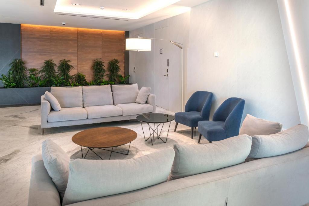 Mūsdienīgs viesnīcas vestibilis ar gaišām mēbelēm, tostarp pelēku dīvānu un zilu krēslu komplektu, papildināts ar dekoratīvām telpaugiem un elegantu kafijas galdiņu.