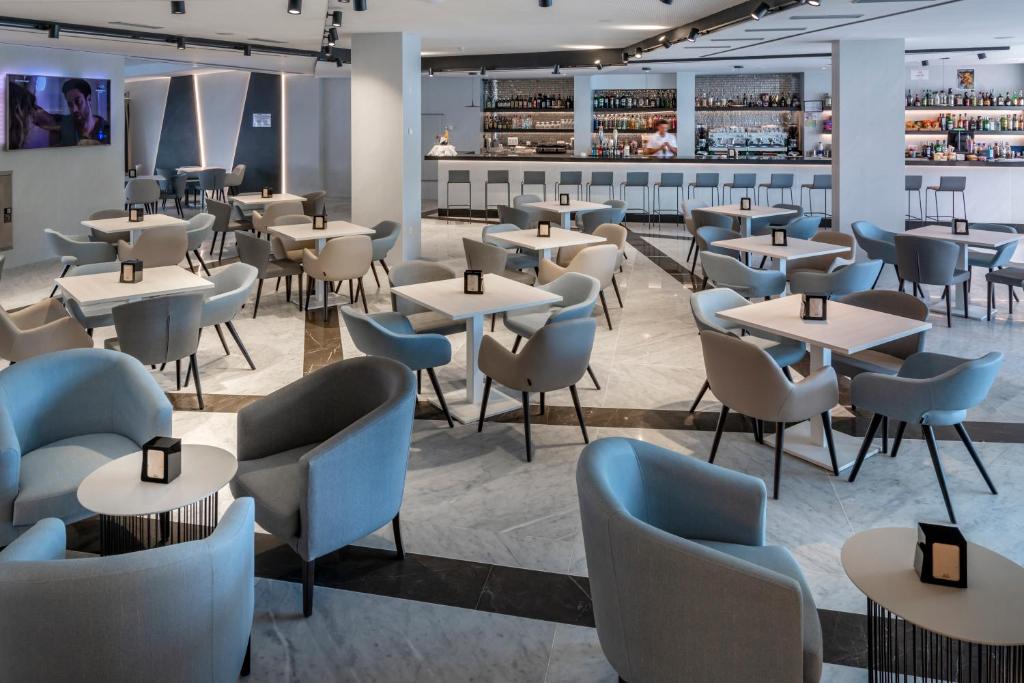 Mūsdienīgs restorāns ar stilīgām pelēkām krēsliem un galdiem, kas izveidots relaksējošai atmosfērai. Fons ar bāru un televizoru, kas piešķir dinamiku telpai.