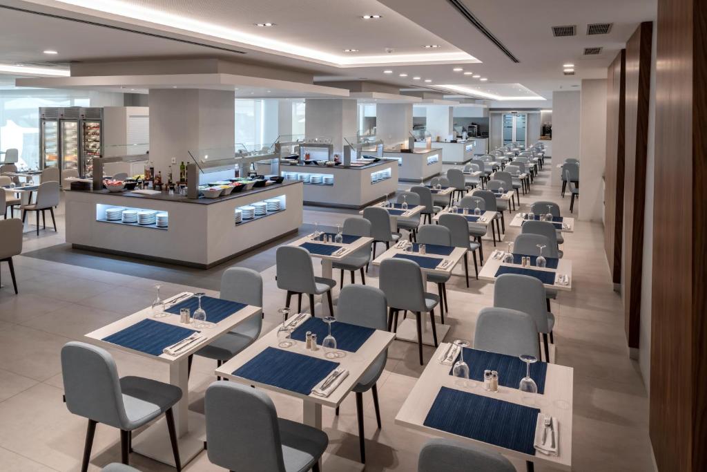 Mūsdienīgs restorāns ar gaišām telpām un elegantiem galdiem, kas sakārtoti ar ziliem galdautiem. Redzama bufete un dzērienu stends, radot viesmīlīgu atmosfēru.