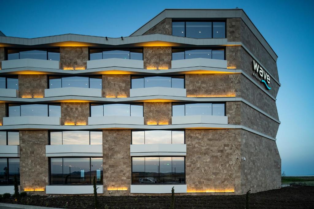 Mūsdienīgas viesnīcas ēka ar akmens apdari un plašiem logiem, kas izceļas ar modernu dizainu un apgaismojumu. Viesnīcas nosaukums "Wave" ir redzams uz fasādes.