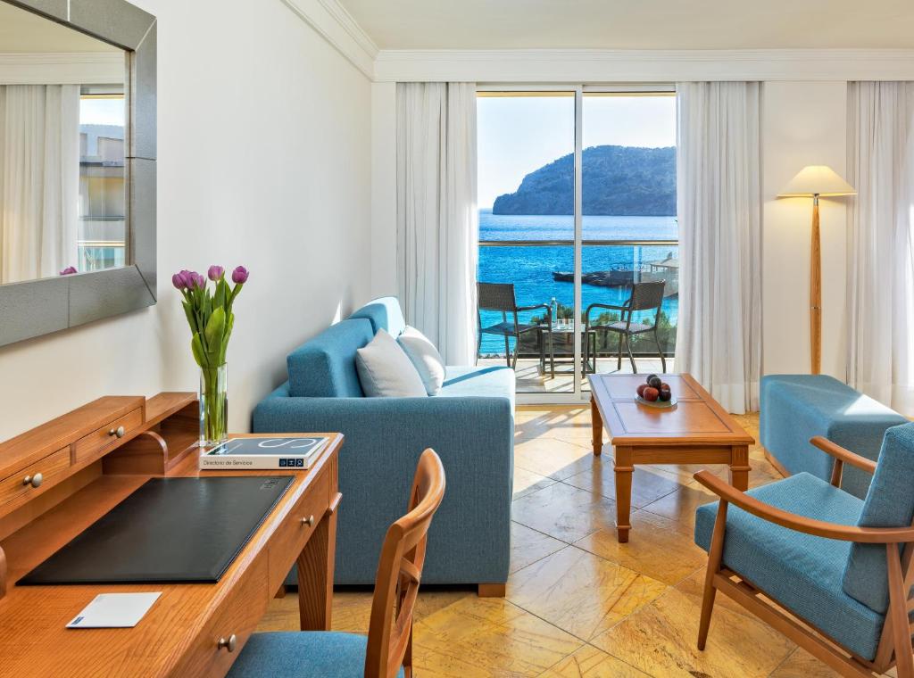 Mūsdienīga viesnīcas numura interjers ar skatu uz jūru, atpūtas zonu un rakstāmgaldu. Tuvumā redzami ziedi un ērtas mēbeles.