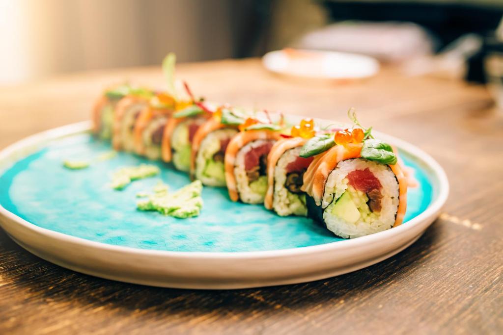 Svaigu sushi ruļļu šķīvis ar krāsainām sastāvdaļām un avokado, pasniegts uz gaiši zila šķīvja.