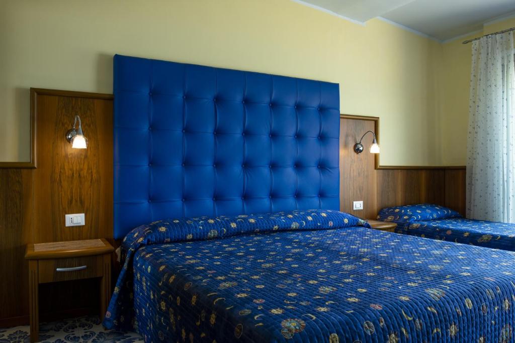 Mūsdienīgs guļamistabas interjers ar zilu gultas galvu un ziedu rakstu gultas veļu. Telpā ir divas gultas un pievienotas lampas, radot ērtu un relaksējošu atmosfēru.