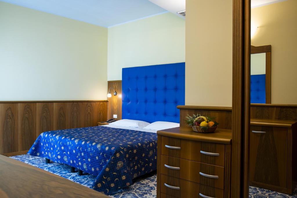 Mūsdienīgs viesnīcas numurs ar zilu gultas pārklāju un koka mēbelēm, kas rada siltu atmosfēru. Numurā redzama arī spoguļa un augļu groza klātbūtne.