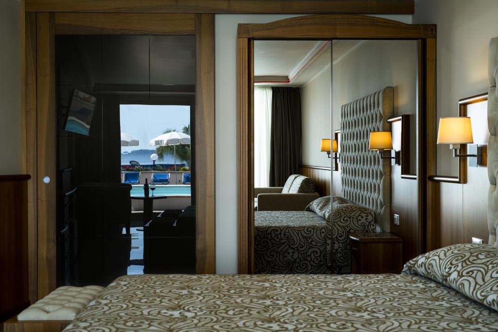 Mūsdienīgs viesnīcas numurs ar elegantu interjeru, kurā redzama gulta ar rakstainu gultasveļu un atspoguļojums, kas rāda skatu uz baseinu un terasi ar saulessargiem.