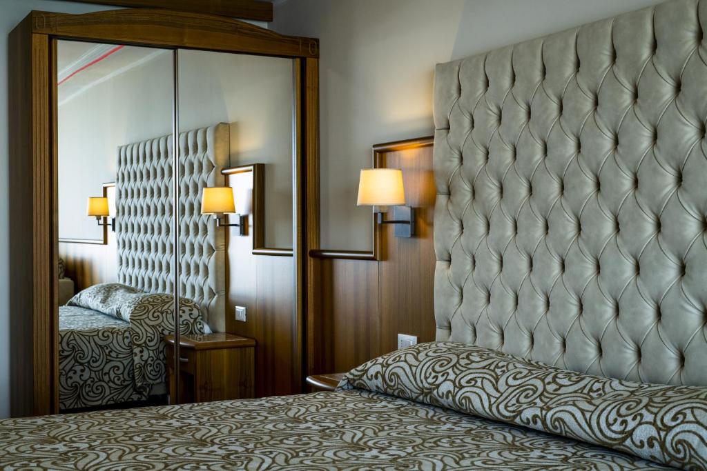 Mūsdienīgas guļamistabas interjers ar izsmalcinātu gultas galvgalu un spoguli, kas atspoguļo elegantu dizainu un komfortu. Sienas lampas piešķir siltumu un atmosfēru.