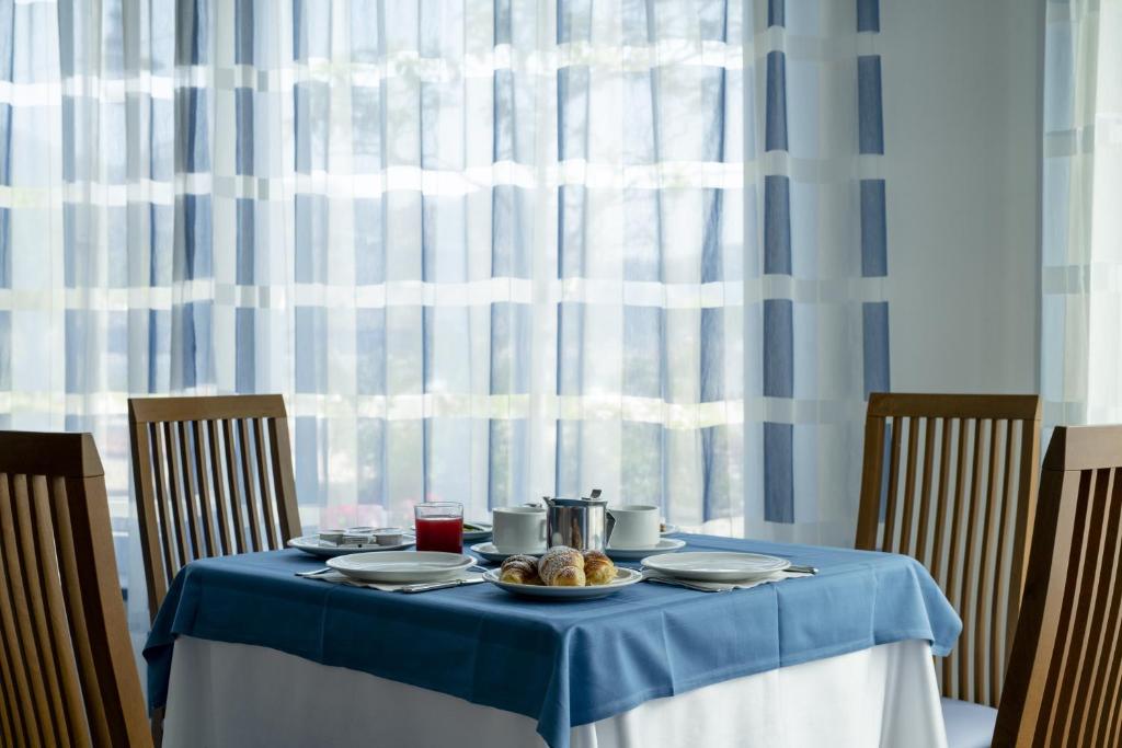 Eleganta brokastu galda uzstādīšana ar zilu galdautu, kafijas un sulas traukiem, kā arī maizītēm, fonā redzami gaiši, svītraini aizkari.