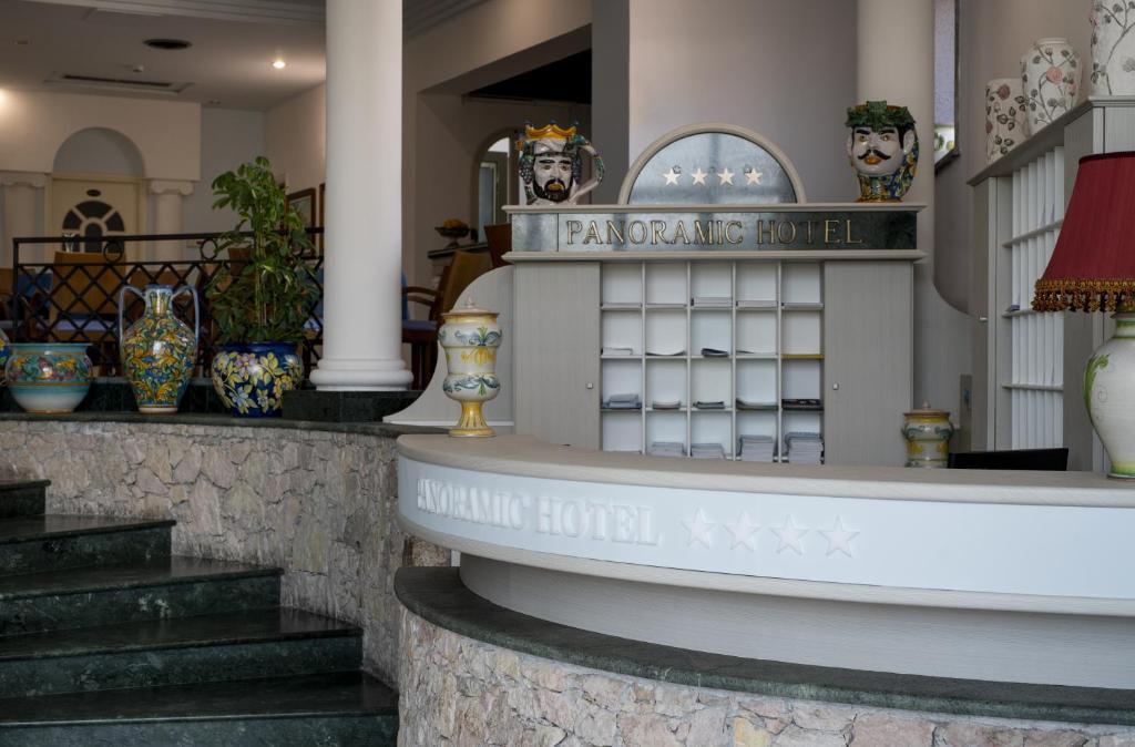 Skaists viesnīcas reģistratūras interjers ar dekoratīviem traukiem un augu elementiem, kas rada viesmīlīgu atmosfēru. Viesnīcas nosaukums "Panoramic Hotel" ir izcelts uz reģistratūras letes.