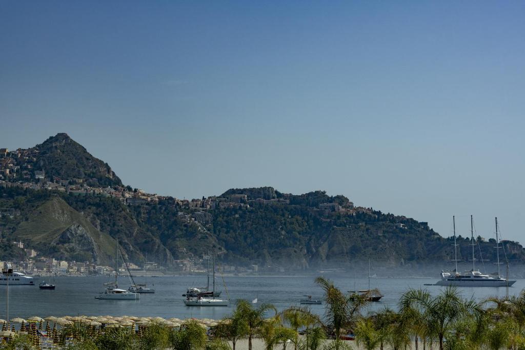 Skats uz kalnainu piekrasti ar kuģiem un jūru, fonā redzama pilsēta. Jūrā plūst daži jahti, bet krastā ir palmas un pludmales sauļošanās krēsli.