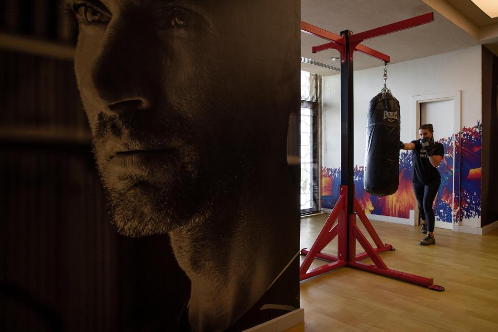 Treniņu telpa ar boksa maisu un sportistu, kas trenējas. Sienā redzams melnbalts attēls ar vīrieša seju, kas piešķir telpai dinamisku atmosfēru.