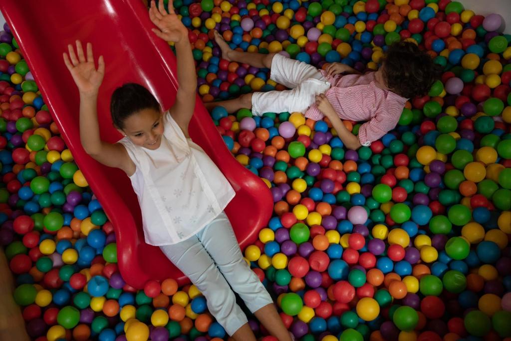 Bērni spēlējas krāsainu bumbiņu baseinā, viena meitene slīd pa sarkano slaidu, kamēr otrs bērns atpūšas bumbiņu vidū.