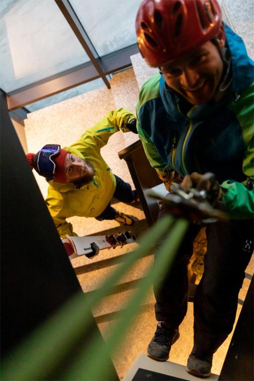 Divi kalnu kāpēji kāpj pa kāpnēm, tērpušies krāsainās sporta drēbēs un ķiverēs, ar ekipējumu rokās. Gaisma ir maiga, radot dinamisku un piedzīvojumu pilnu atmosfēru.