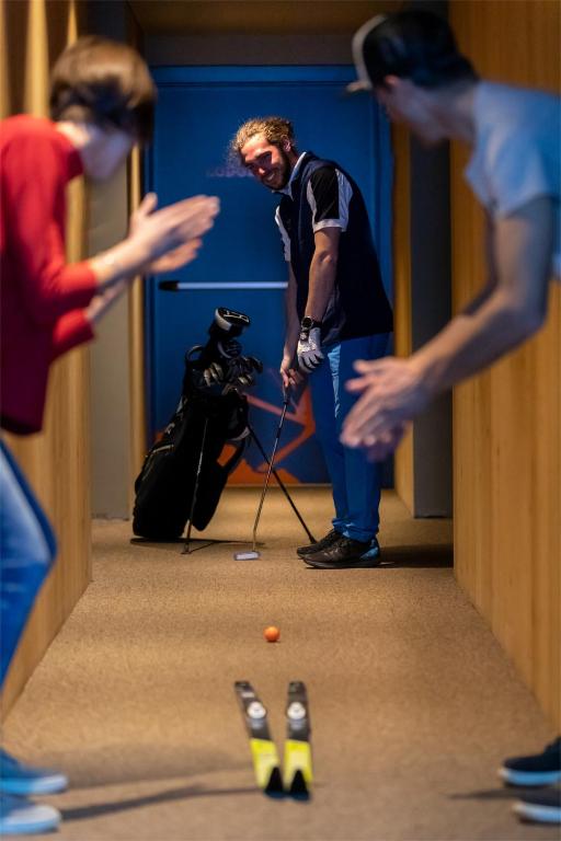 Divi cilvēki aplaudē, kamēr trešais vīrietis spēlē golfa putteru šaurā koridorā, kas ir aprīkots ar golfa ekipējumu. Golfā tiek izmantota oranža bumbiņa, un priekšā ir slēpošanas dēļu pāris.