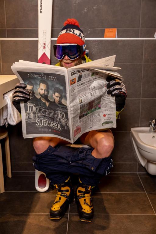 Vīrietis sēž uz poda, tērpies slēpošanas apģērbā un dzeltenās slēpošanas zābakos, lasot avīzi. Fons ir moderns, ar flīzēm un slēpēm.