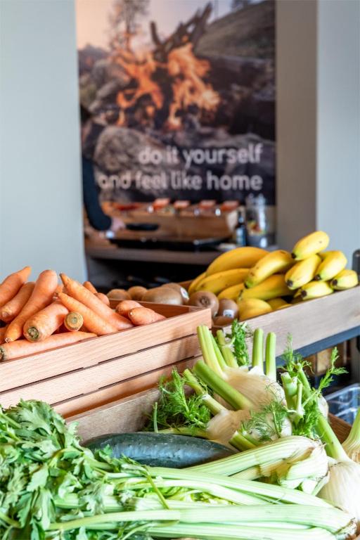 Dārzeņu stends ar burkāniem, gurķiem un selerijām, kā arī banāniem fonā ar aicinājumu "dariet paši un jūtieties kā mājās".