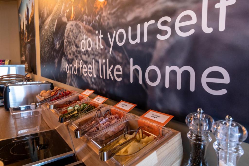 Pašapkalpošanās stūra attēls ar dažādām sastāvdaļām, piemēram, dārzeņiem un gaļu, kas novietotas caurspīdīgās traukos. Fons satur uzrakstu "do it yourself and feel like home".