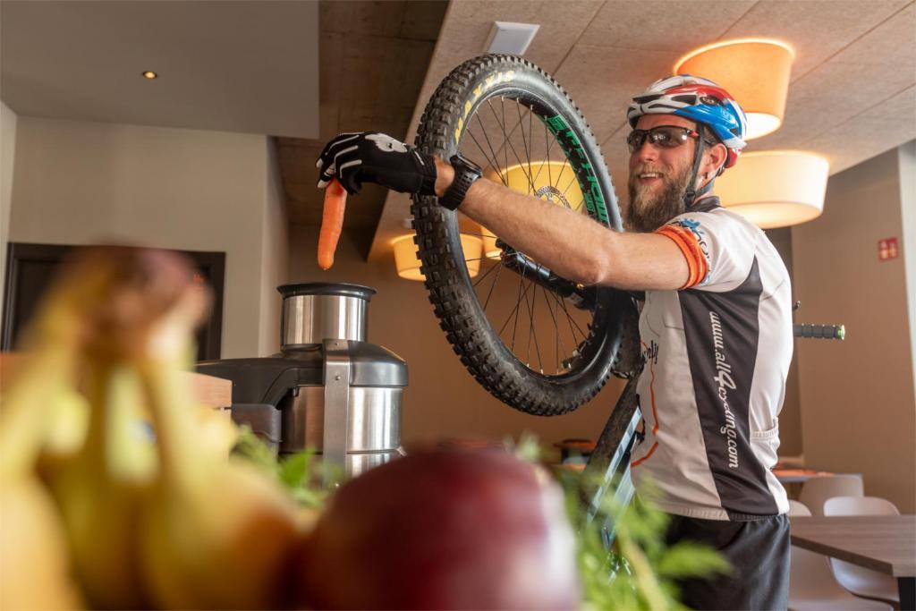 Cilvēks ar velosipēdu, turot to augšā, kamēr viņš pieliek burkānu pie sulas spiedes. Fons ir kafejnīca ar augļiem un sulas iekārtu.