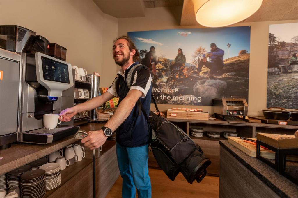 Jaunais vīrietis, kas smaida, gatavo kafiju ar automātu, turot grozu un ar mugursomu uz pleca. Fons rāda dažādus kafijas traukus un ainavu ar cilvēkiem, kas bauda dabu.