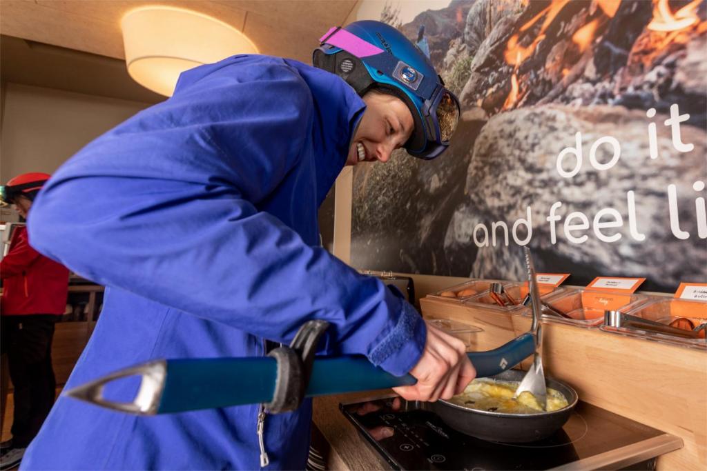 Jauns vārds tiek gatavots virtuvē, kur sieviete ar ķiveri un sniega apģērbu smaida, izmantojot pannas un virtuves rīkus. Fons ir dekorēts ar mudinājumu baudīt dzīvi.