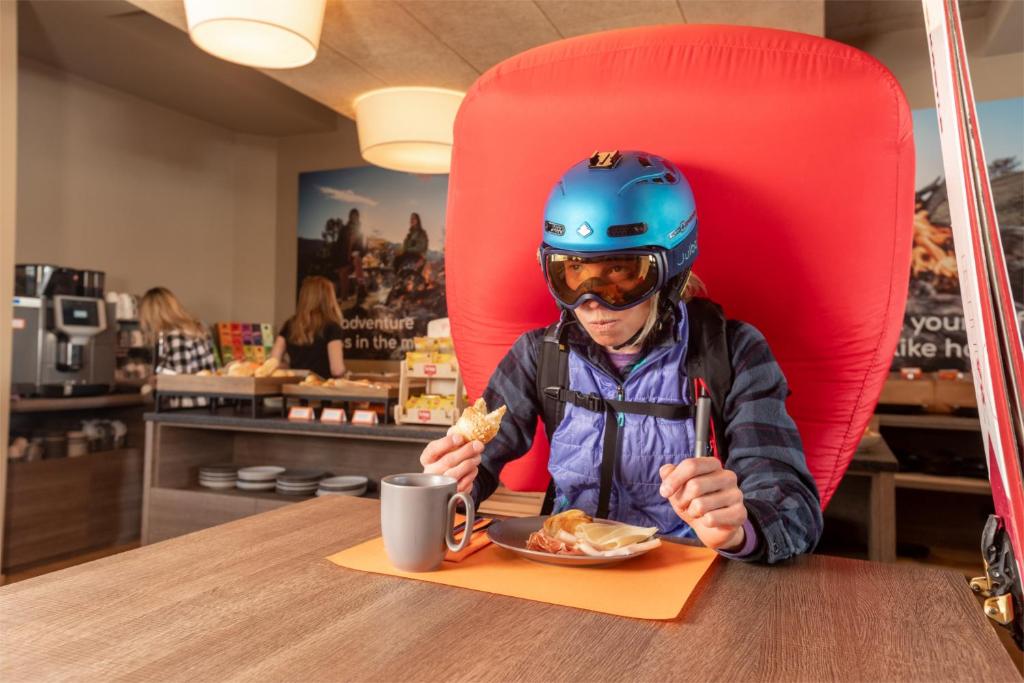 Sniega sporta entuziasts, tērpies slēpošanas ekipējumā, ēd brokastis kafejnīcā, sēžot aiz lielas sarkanas aizsargbarjeras. Galda priekšplānā ir kafijas krūze un šķīvis ar ēdienu.