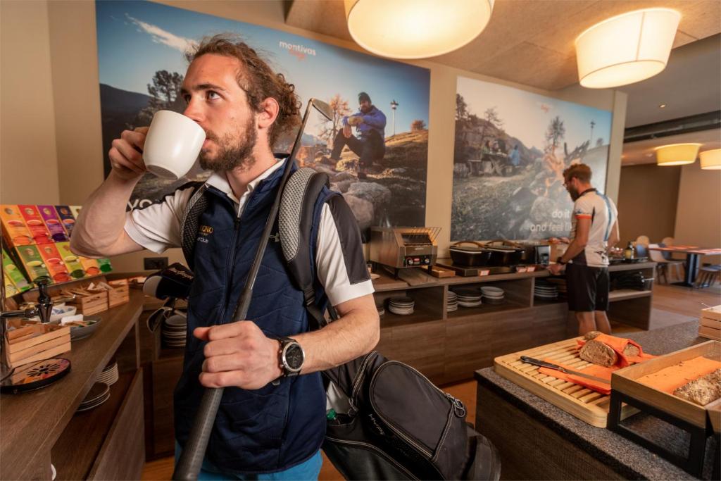 Vīrietis dzer kafiju, stāvot pie brokastu galda, kur ir dažādi ēdieni un dzērieni. Fons parāda kalnu ainavas ar citiem cilvēkiem.