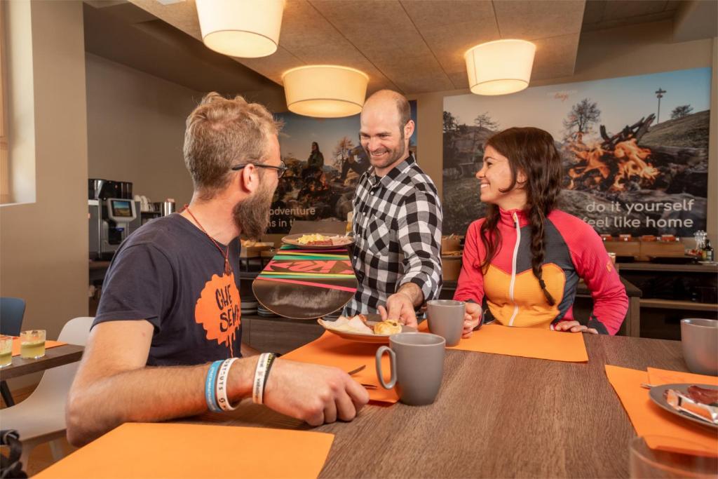 Sociāla vide, kurā cilvēki baudā brokastis, ar viesmīli, kas pasniedz ēdienu. Interjerā redzami silti toņi un moderns dizains, radot mājīgu atmosfēru.