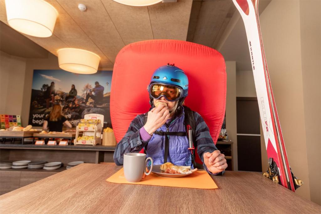 Ski entuziasts, ģērbies pilnā ekipējumā, ēd brokastis kafejnīcā, sēžot pie galda ar skatu uz dažādiem ēdieniem. Fons iekļauj slēpošanas aprīkojumu un viesmīlīgu atmosfēru.