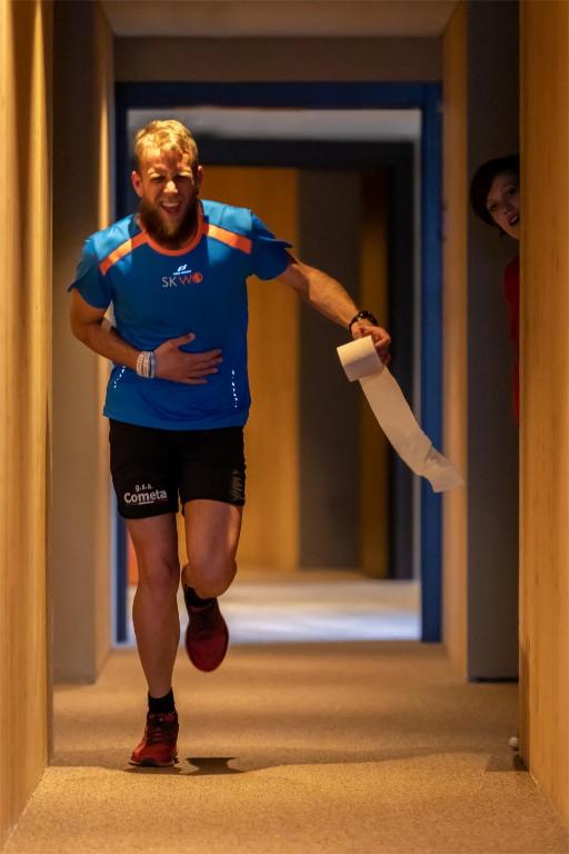 Vīrietis sportiskā tērpā skrien pa garu koridoru, turēdams rokās papīra ruļļu, kamēr aiz viņa izspiegā sieviete.