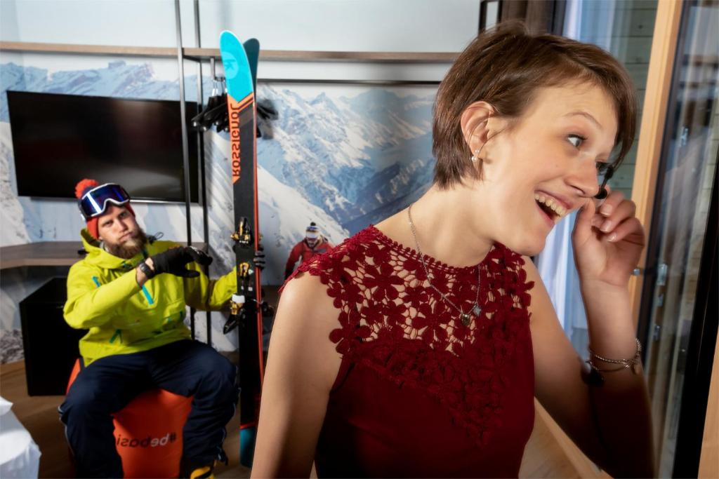 Sniega sporta entuziasts un sieviete ar priecīgu izteiksmi, stāvot mūsdienīgā telpā ar kalnu ainavu fona. Vīrietis sēž uz krēsla ar slēpēm rokās, kamēr sieviete izskatās pārsteigta un laimīga.