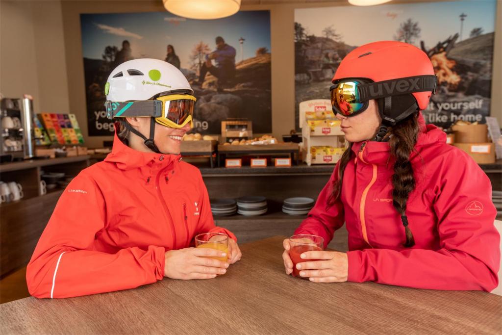 Divas slēpotājas, tērptas sarkanos apģērbos un aizsargbrillēs, sēž kafejnīcā, baudot dzērienus un sarunājoties. Fons veido mājīgu atmosfēru ar kalnu ainavām.