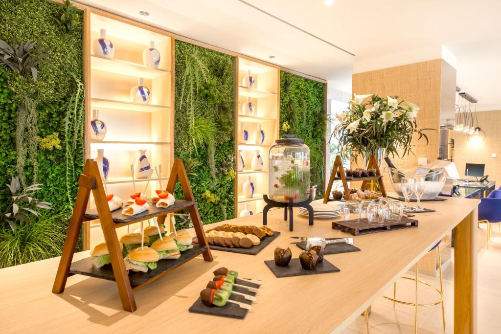 Elegantā bufete ar gardumiem, ieskaitot mini burgerus un saldumus, ir izvietota uz koka galda, ko rotā zaļie augi un dekoratīvi trauki. Interjers ir moderns un viesmīlīgs.