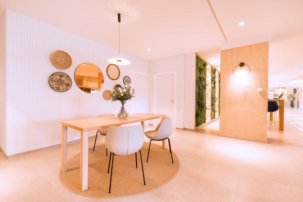 Moderns, gaišs ēdamistabas interjers ar koka galdu, baltām krēsliem un dekoratīvām sienas platekrāsām. Telpā ir ziedu pušķis un dabiski elementi, piešķirot siltumu un mājīgumu.