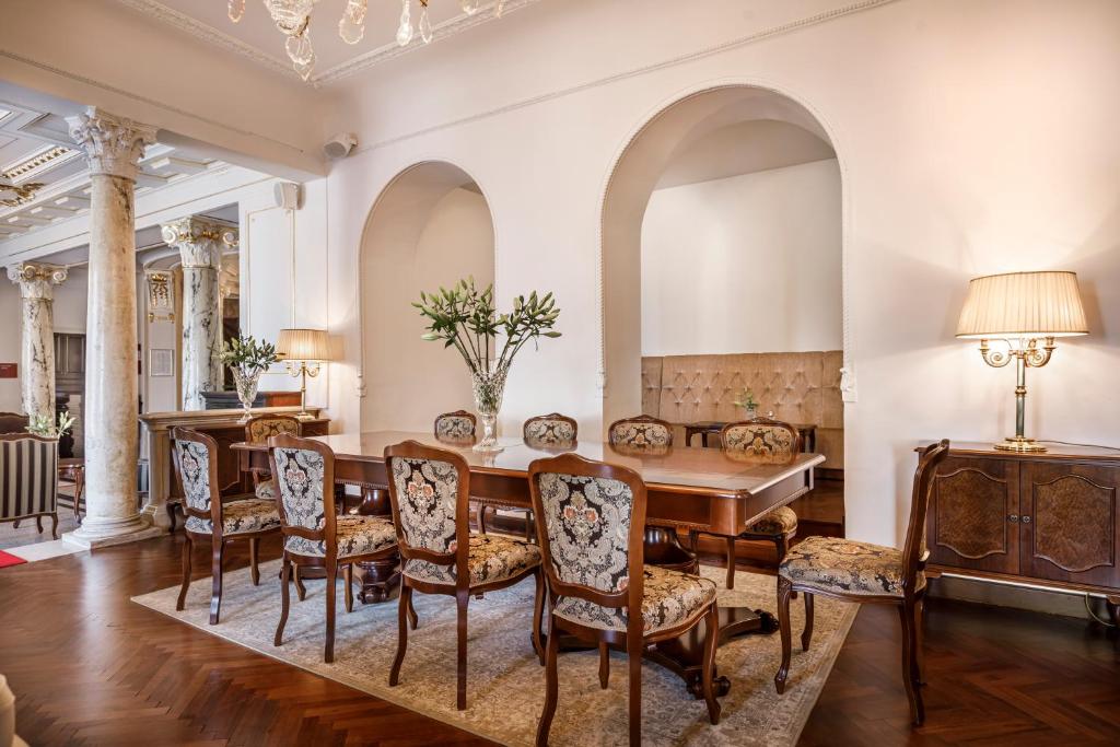 Elegants viesistabas interjers ar koka galdu un greznām krēslām, dekorēts ar ziediem un lampām, radot siltu atmosfēru. Stūri rotā kolonnas un arku, piešķirot telpai klasisku šarmu.