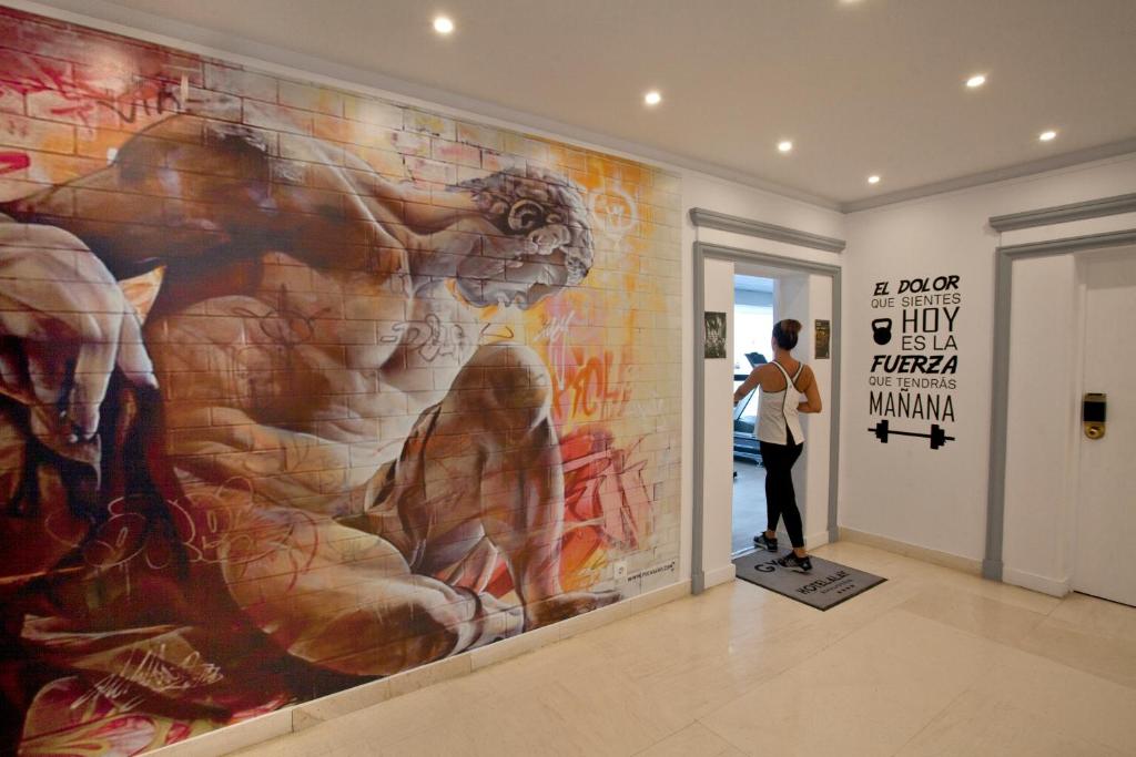 Sienas glezna ar muskuļotu figūru un grafiti elementiem sporta zālē, blakus ir motivējošs citāts par spēku un sāpēm. Iekšējās telpas dizains veicina dinamisku un uzmundrinošu vidi.