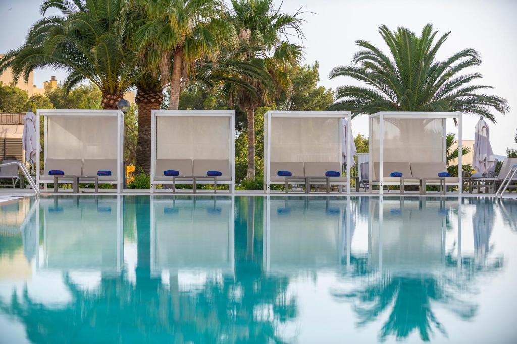 Baseins ar palmām un atpūtas zonām, kas nodrošina relaksējošu atmosfēru. Ūdens virsma atspoguļo apkārtējos elementus.