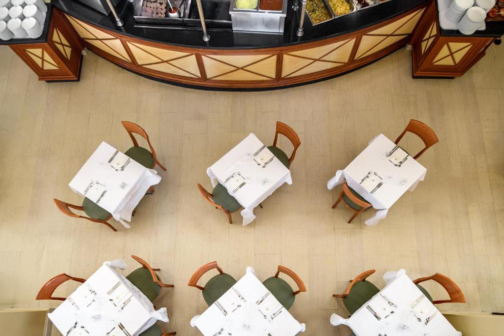 Restorāna interjers ar baltām galdiem un koka krēsliem, redzams no augšas. Galdi ir klāti ar galda piederumiem, un fonā ir bārs ar dažādiem ēdieniem.