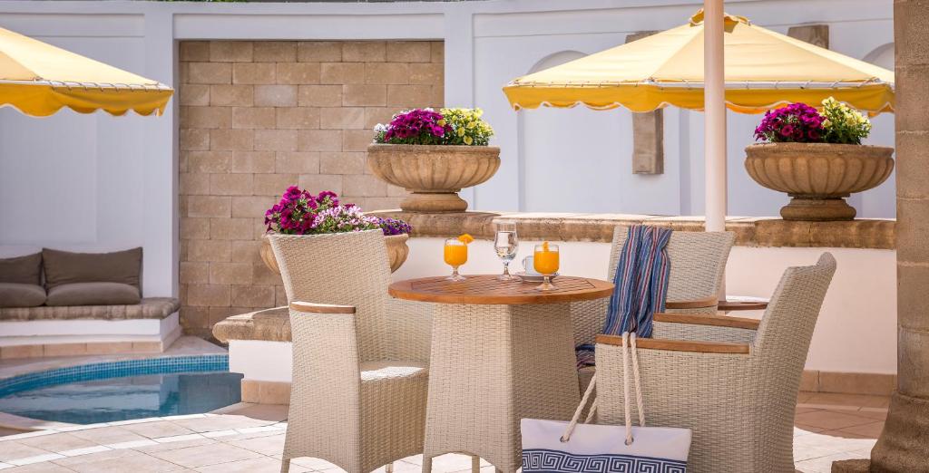 Skaista atpūtas zona ar baltiem krēsliem un koka galdu, ap kuru ir dzērieni un ziedu kompozīcijas, ar dzeltenām saules segām un peldbaseinu fonā.