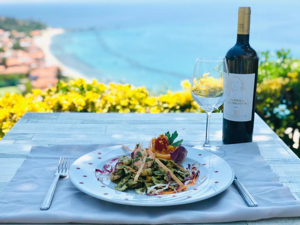 Garšīgs ēdiens ar makaronu šķēlītēm un dārzeņiem, pasniegts uz galda ar vīna pudeli un glāzi, fons ietver jūras ainavu un ziedošus krūmus.