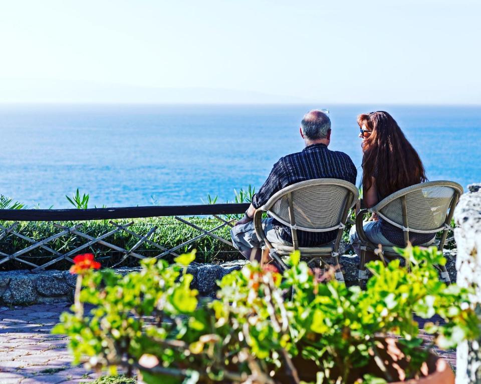 Pāris sēž krēslos, raugoties uz jūru, kamēr saulaina diena apgaismo ainavu. Prieks par dabu un mierīgu brīdi kopā.