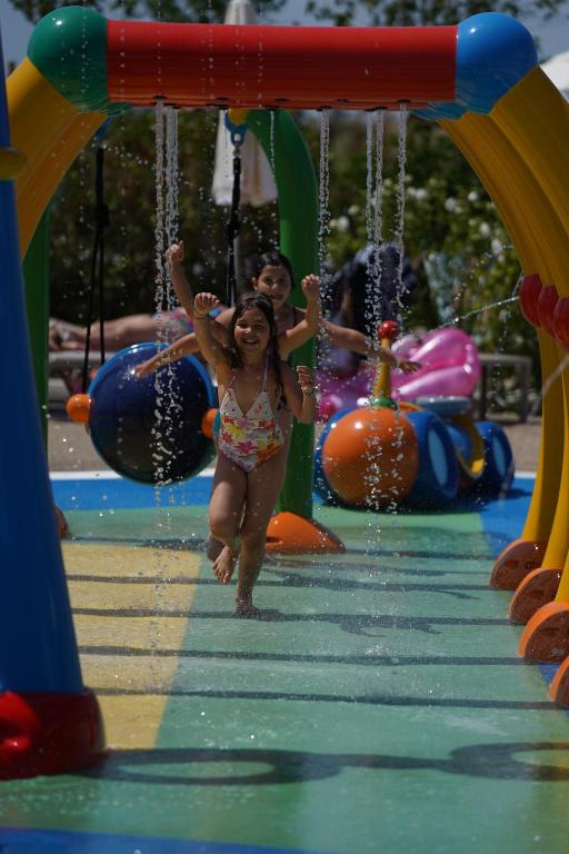 Bērni spēlējas ūdens atrakciju parkā, skrienot cauri ūdens strūklām. Krāsainas ūdens atrakcijas un rotaļlietas veido priecīgu atmosfēru.