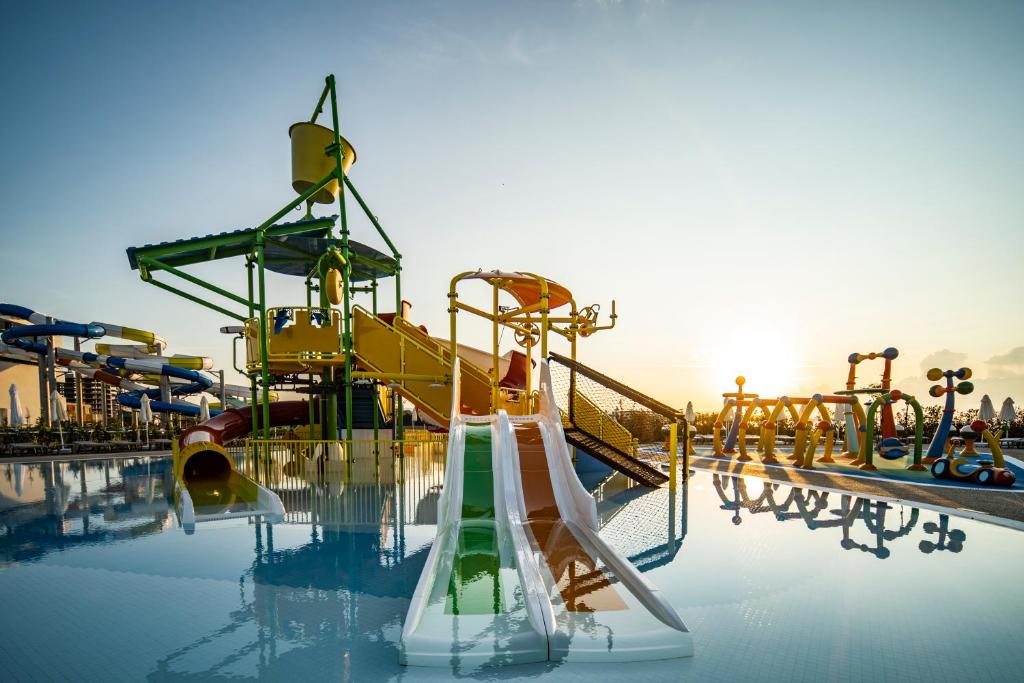 Ūdens atrakciju parks ar krāsainām ūdens slīdēm un rotaļu elementiem, saulrieta gaismā. Ūdens virsma atspoguļo struktūras un debesis.