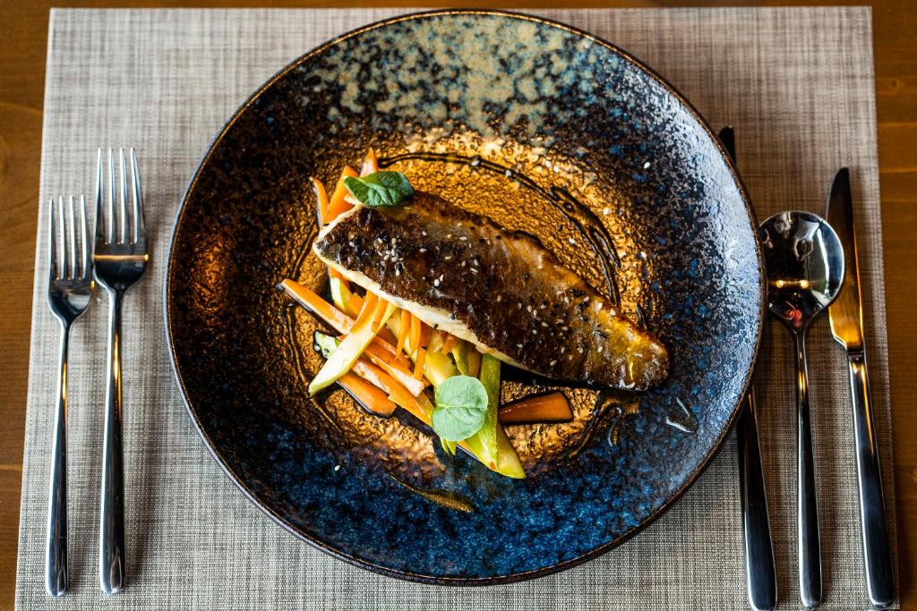 Garšaugu zivs fileja uz dārzeņu gultas, pasniegta elegantā melnā šķīvī ar sudraba galda piederumiem.