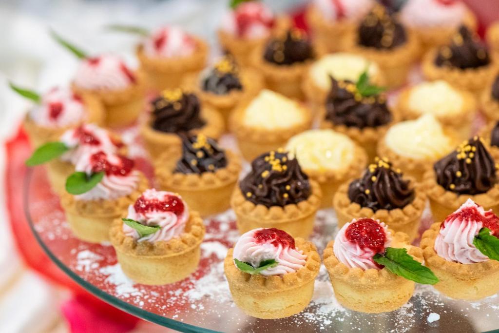 Saldumu kārumi uz stikla šķīvja, tostarp mini tortes ar krēmīgu virskārtu un augļu dekorācijām. Izskatās krāsaini un pievilcīgi, ideāli piemēroti svinībām.