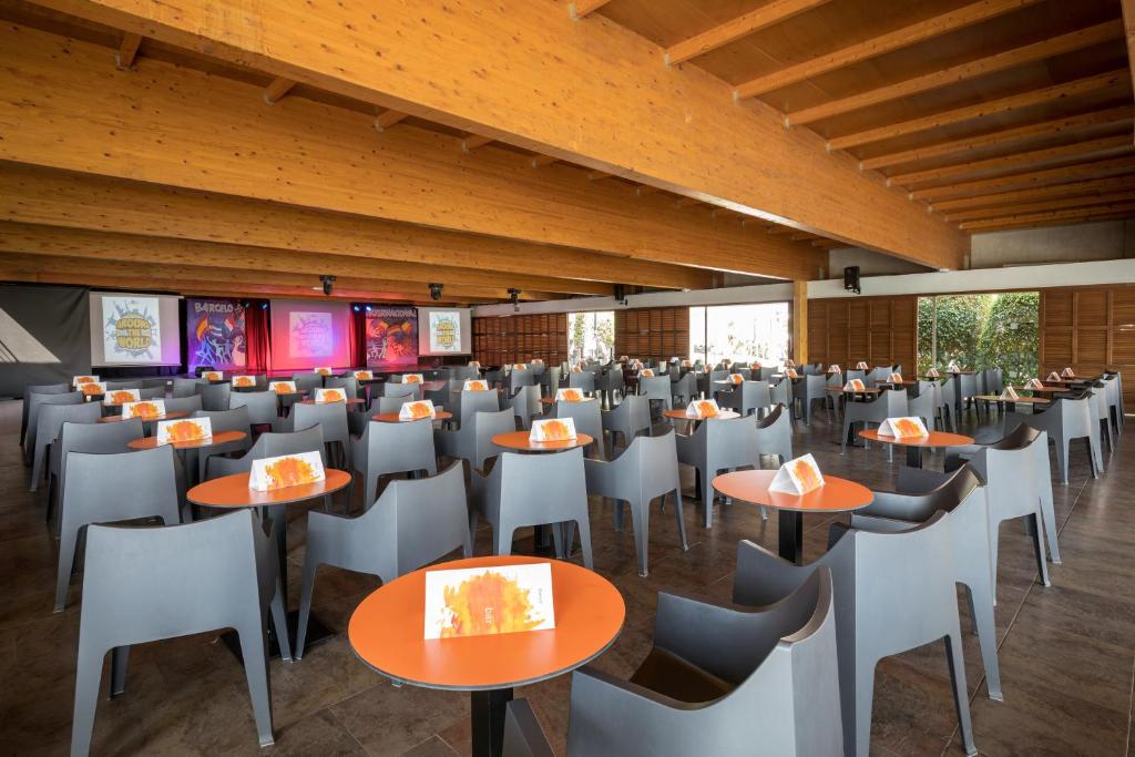 Liels un moderns pasākumu zāles interjers ar melniem krēsliem un oranžām galdiņām, gatavs viesu uzņemšanai. Aizmugurē redzami ekrāni ar spilgtām reklāmām.