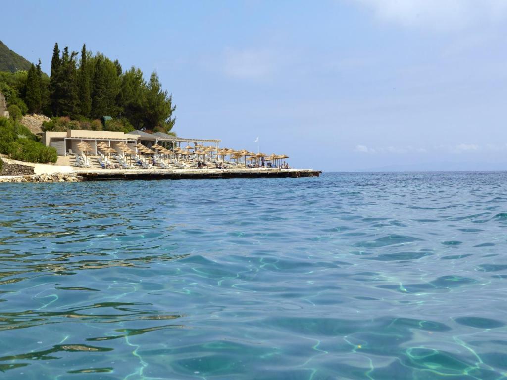 Pludmales zona ar sauļošanās krēsliem un saulessargiem pie skaidra ūdens. Skaista ainava ar zaļām koku grupām un kalnu fona.